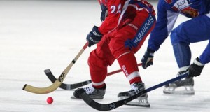 2 bandy ball players one red one blue following orange ball 3 hockey sticks basic equipment hockey sticks orange round bandy ball